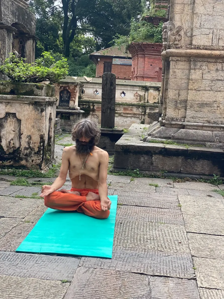 Yogi Ji, Ashok Sharma Bhatta, seated in lotus on a mat in one of the old stone courtyards at Pashupatinath, carved shrines and moss-covered walls around him