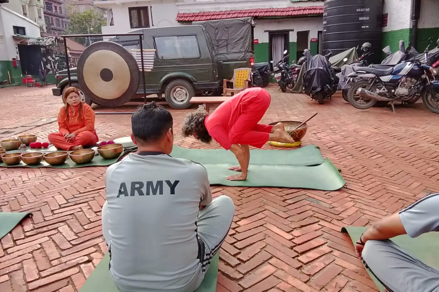 An outdoor courtyard session with personnel seated on mats, a gong on its stand and singing bowls laid out in front